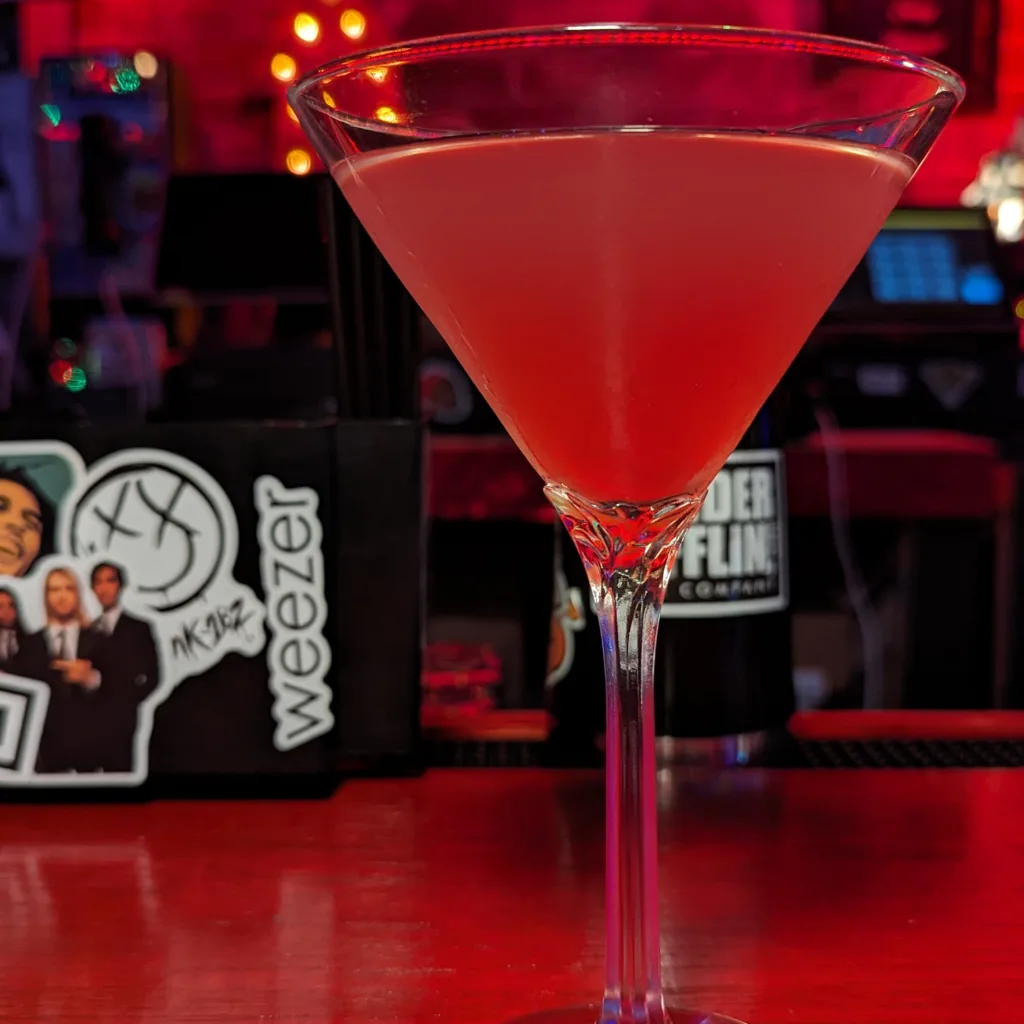 A pink cocktail in a martini glass on a bar counter with dim lighting and a backdrop featuring a weezer poster.